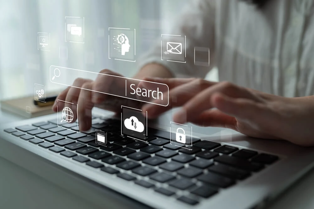Hände tippen auf einer Laptop-Tastatur, umgeben von digitalen Icons einer Suchmaschine, darunter eine Suchleiste und ein Globus-Symbol