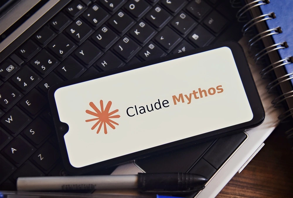 Smartphone mit dem Claude-Mythos-Logo auf dem Display, platziert auf einer Laptop-Tastatur neben einem Notizbuch und einem Stift.