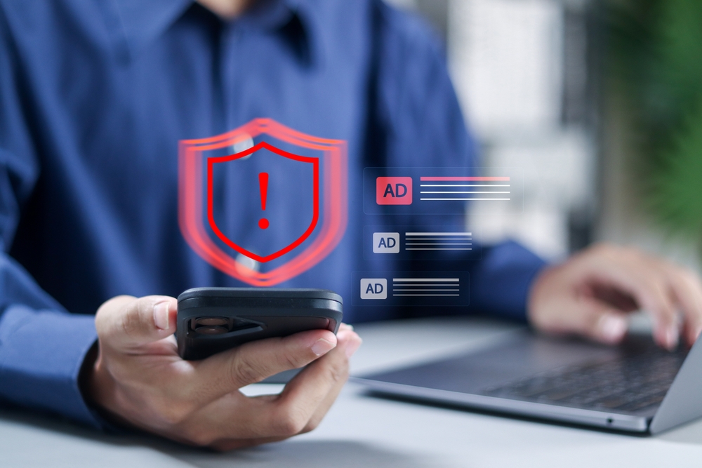 Person hält Smartphone mit Sicherheitsschild und Ausrufezeichen vor Laptop, im Hintergrund Anzeigen-Icons